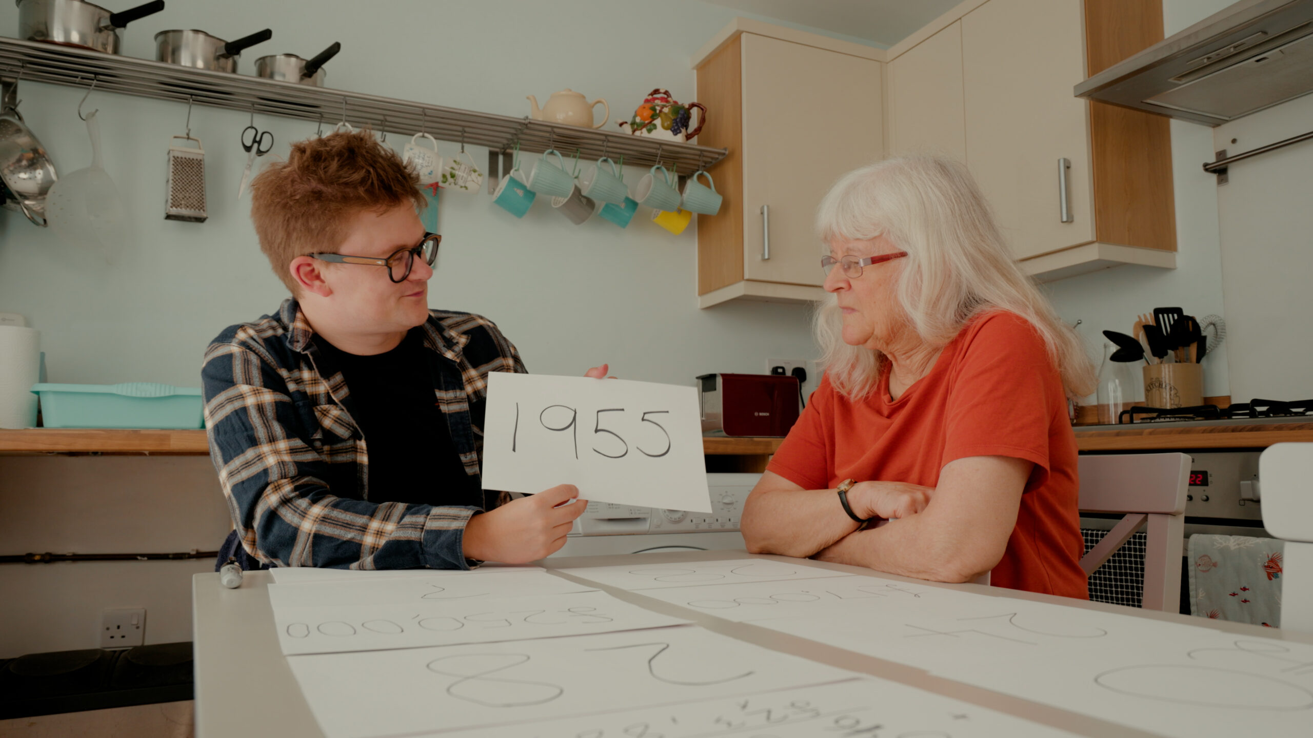 Tom Nicholas and an elderly woman sit at a kitchen table. Tom holds up a sheet of paper that has '1955' written on it.